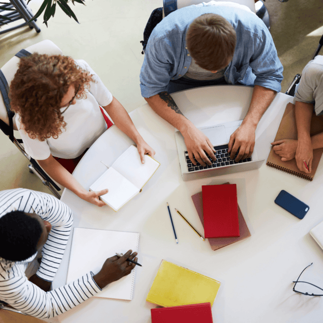 Head shot view of 4 people sitting at a round table learning together using pen and paper and laptop